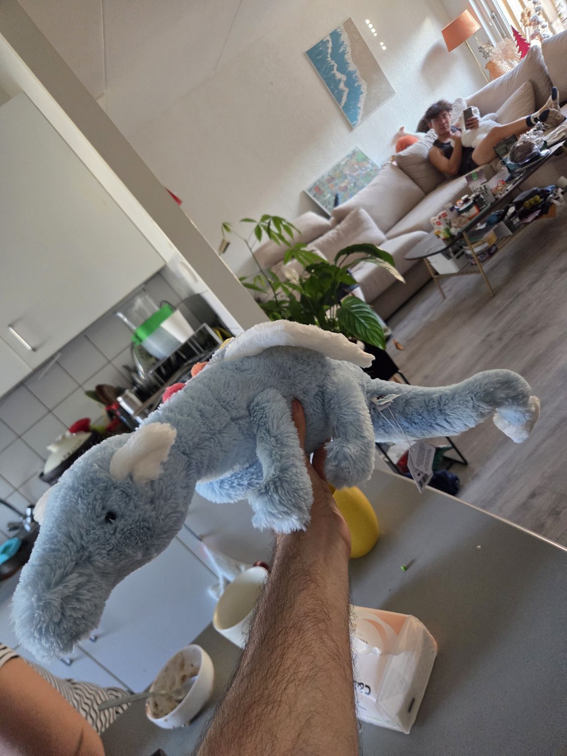 A pale blue-grey plush elephant with cream ears held up at arm's length in a sunlit apartment, kitchen counter in front, sofa and houseplant behind.